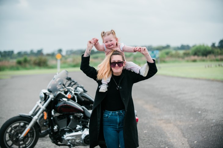 Kavana Jane holding her daughter Harley-Jean on her shoulders in front of a motorbike.