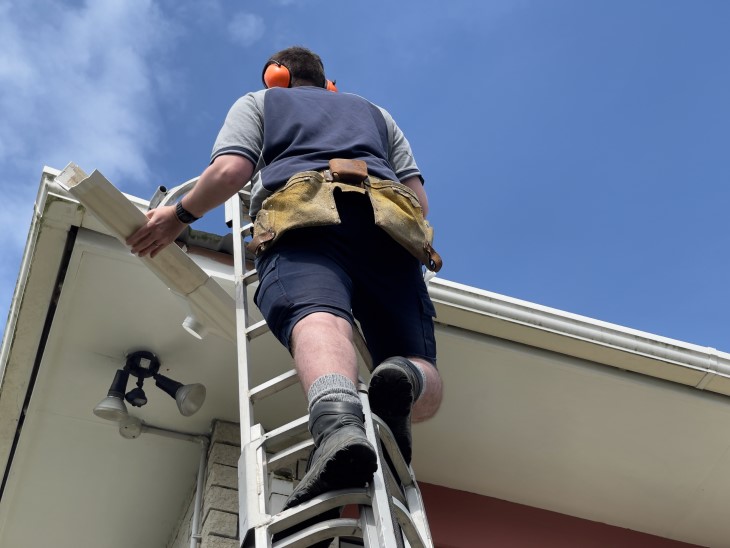 A photo taken from below showing a man on a ladder fixing the guttering on a roof.