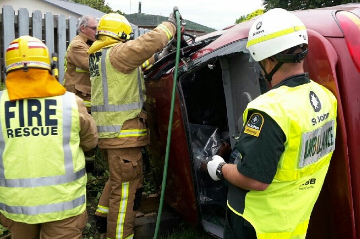 Derek stands by a crashed car on it's side in his paramedic gear while firefighters cut the car open