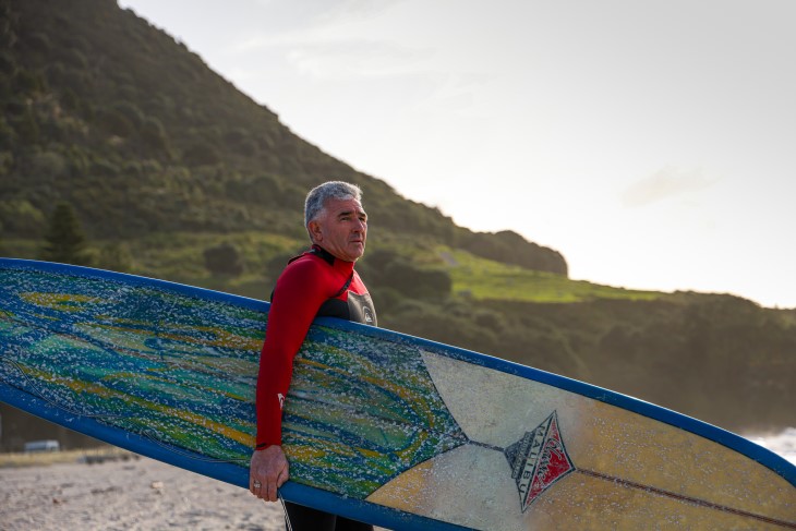 Michael holding a surfboard on the sandy beach shore.