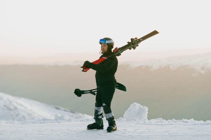 Paralympian Adam Hall standing at the top of a snow-covered mountain holding his skis./
