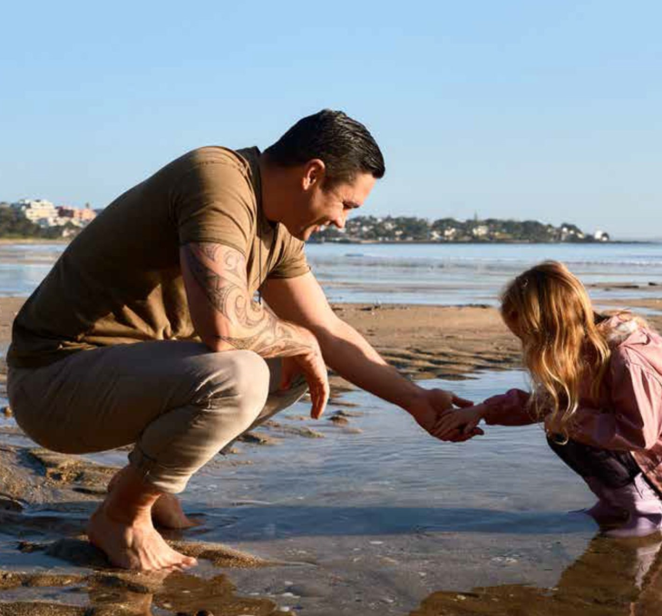 A father kneeling down with his young daughter to look at something in the shallow water at a beach.