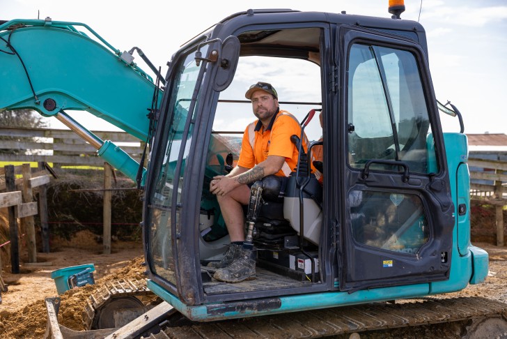Cory McKinnon wearing his prosthetic leg sitting in the cabin of a digger on a construction site.