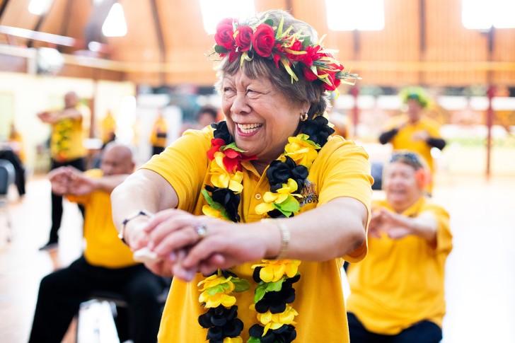 An older Pasifika woman doing exercises during a group class in a gymnasium.
