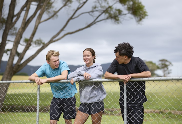 Three young people leaning on a fence at a sports field.