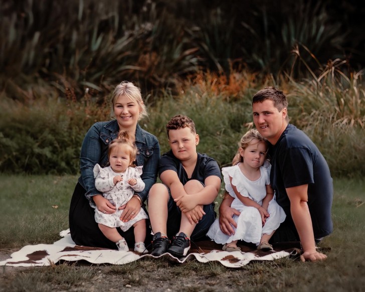 A family photo showing Mark Julian sitting down on a rug on the grass with wife Emily and their three children.