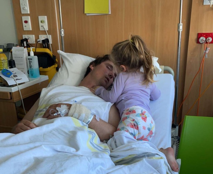 Mark Julian lying in a hospital bed with his young daughter hugging him.