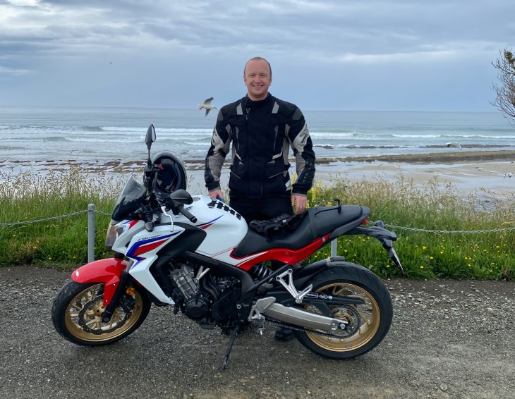 Nick Grant standing behind his motorbike in front of a beach.