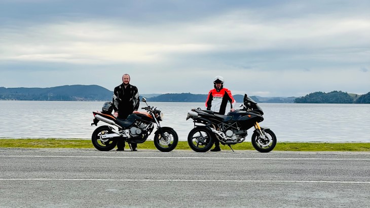 Nick Grant standing with his father and their motorbikes on a road beside the ocean.