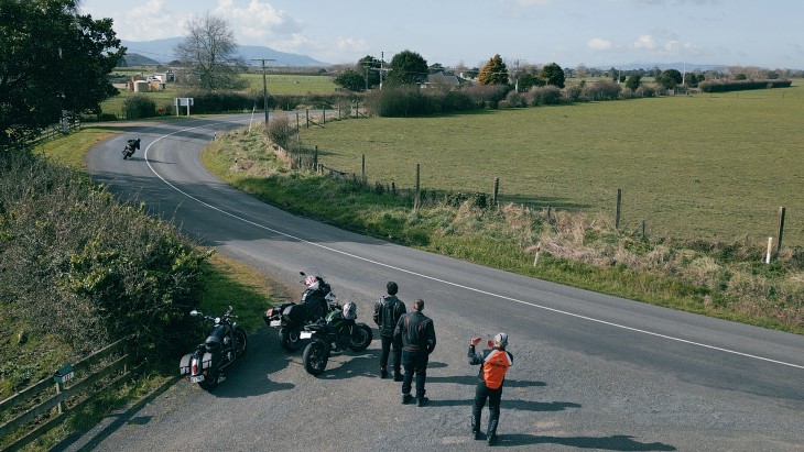 A pair of motorcyclists standing next to their bikes and watching another motorcyclist riding on a corner while a Ride Forever instructor teaches them.