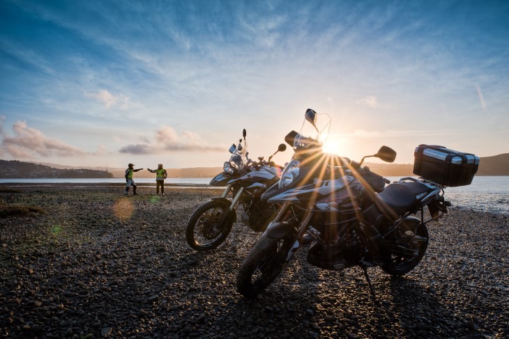 Two motorbikes on the beach as the sun sets with their owners standing in the background.